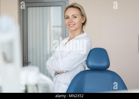 Ritratto di bellissima femmina dentista in posa di office della moderna clinica dentale, tranquillamente in piedi con le braccia incrociate e guardando la fotocamera Foto Stock