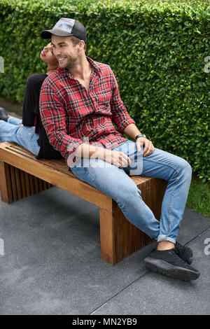 Carino interracial paio di sedersi con le loro spalle per ogni altro sul banco di legno sul verde sfondo boccole all'esterno. Essi sono sempre sorridenti. Ragazzo bianco e bl Foto Stock