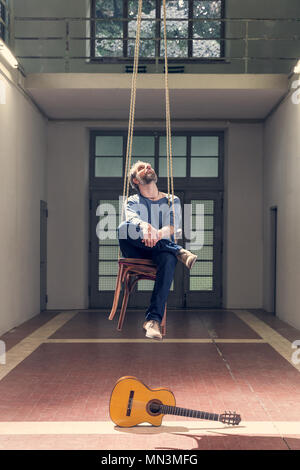 Uomo seduto su una sedia pensile e una chitarra sul pavimento, interno Foto Stock