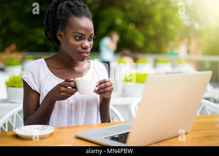 Affascinante African American donna imprenditore è seduto al ristorante di strada e avente online in remoto sessione video con i colleghi utilizzando il computer portatile, caldo Foto Stock