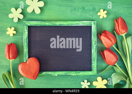 Vacanze di primavera sullo sfondo: blackboard incorniciato con tulipani rossi, cuore di legno e fiori su legno verde, lo spazio per il tuo testo Foto Stock