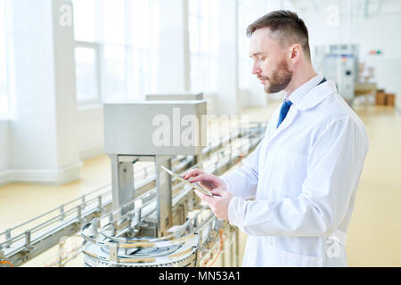 Vista di profilo di fiducioso barbuto operatore della macchina indossando camice bianco con tavoletta digitale mentre avvolto fino al lavoro moderno reparto di produzione di Foto Stock