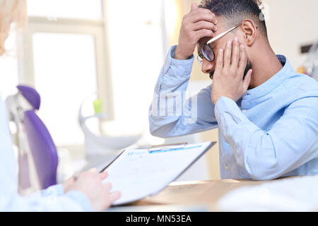 Vista laterale ritratto di preoccupato uomo mediorientale che soffrono di mal di denti e azienda guancia gonfia mentre si parla al dottore seduto alla scrivania nella moderna Foto Stock