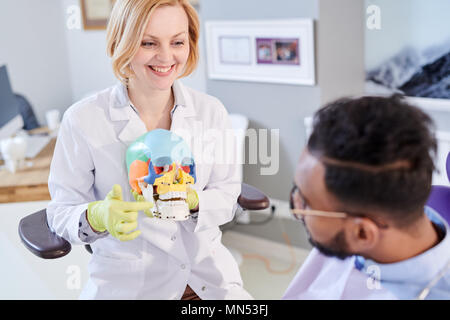 Ritratto di donna sorridente dentista azienda modello di cranio e spiegare i denti in materia di igiene per la giovane uomo seduto in poltrona odontoiatrica Foto Stock