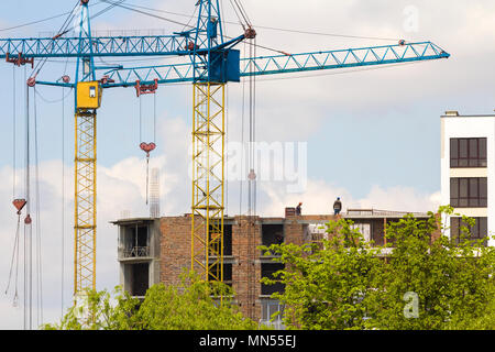 Vista urbano di sagome di due alte Industrial Gru a torre a lavorare alla costruzione di un nuovo edificio di mattoni con i lavoratori di copricapi rigidi su di esso contro la br Foto Stock