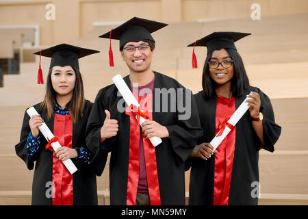 Sorridente diverse nazionalità laureati mantenendo i diplomi in piedi in spacy moderna università in aula. Gli studenti che indossa il nuovo nero e rosso abiti di graduazione. Sentirsi felice e soddisfatto, ridendo. Foto Stock