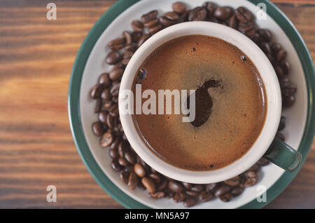 Una tazza di caffè forte con una schiuma su un piattino con i chicchi di caffè su un sfondo di legno Foto Stock