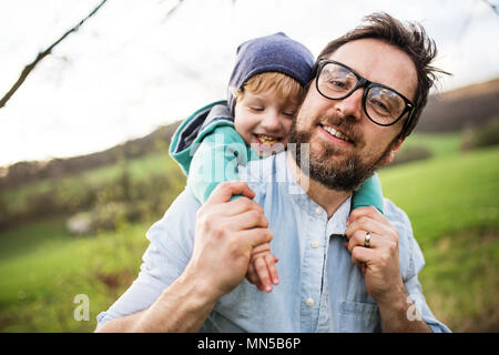 Un padre tiene il suo bimbo figlio sulla schiena fuori nel sole verde primavera la natura. Foto Stock