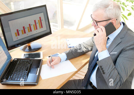 Alta angolazione del senior investment imprenditore indossare tuta mentre seduto alla scrivania in ufficio nella parte anteriore del computer portatile ed effettuare una chiamata. Foto Stock