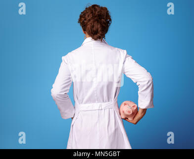 Visto da dietro il medico donna in bianco manto medico tenendo piggybank isolato su blu Foto Stock