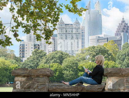 Bella donna seduta sul muro di pietra in Piemonte Park a giocare con il suo telefono cellulare. Atlanta skyline è in background. Foto Stock