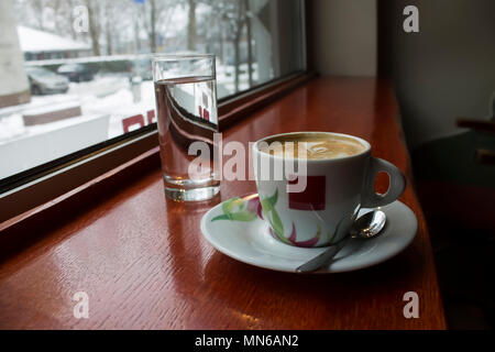 Fresca bruna, caffè con un bel disegno e un bicchiere di acqua su un lungo, rosso, tavolo di legno accanto a Windows nella barra e la neve al di fuori Foto Stock