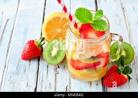 Acqua detox di limone, fragola, kiwi in un bicchiere di vasetto di mason con frutta sparsa su uno sfondo rustico di legno blu Foto Stock