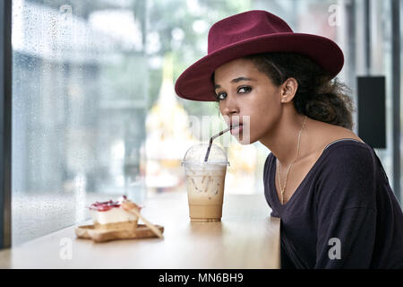 Deliziosa ragazza ricci beve un cocktail in una caffetteria sul background windows. Indossa una dark maniche lunghe con un cappello rosso cremisi e guarda nella telecamera. Foto Stock