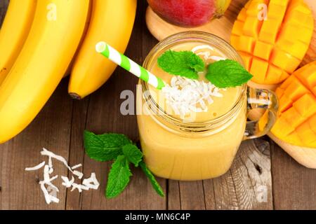 Mango, banana, noce di cocco frullati in una giara di mason vetro, vista aerea su un rustico sfondo legno Foto Stock