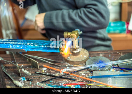 Le mani dell'artigianato uomo che fa un oggetto di vetro presso la fiera di artigianato.produttore di vetro lavorando il vetro fuso con una torcia ardente. In prossimità di fiamme da bl Foto Stock