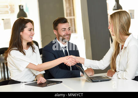 Sorridente coppia giovane stringono le mani con un agente di assicurazione o consulente di investimento. Tre persone che si incontrano in un ufficio di giungere a un accordo Foto Stock