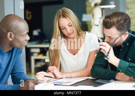 Tre giovani che studiano con la grafica sul tavolo bianco. Bella ragazza bionda, africana e uomini caucasici lavorando insieme di indossare un abbigliamento informale. Multi-gruppo etnico. Foto Stock