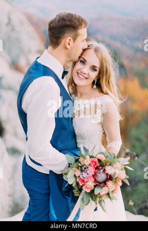 Lo Sposo è baciare la sposa sorridente in fronte al background del ingiallito foresta. Foto Stock