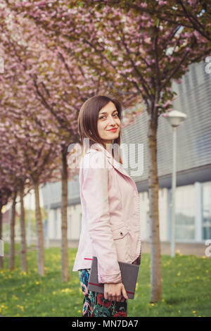 Ritratto di donna su rosa alberi in fiore nella strada della citta'. Giovane e bella signora tenendo il suo notebook di carta vicino edificio moderno Foto Stock