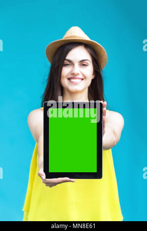 Brunette donna vestita in maglietta gialla e hat detiene il tablet PC con schermo verde prima di sfondo blu, pubblicizzare il concetto di spazio Foto Stock
