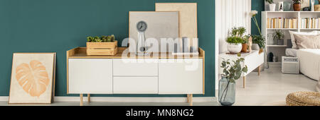 Bianco credenza in legno con dipinti, grigio candele, orologio e di piante fresche in piedi in uno spazio aperto sala interna con pareti verdi, letto e libri Foto Stock