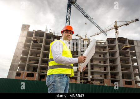 Ritratto di ingegnere maschio in hardhat e sicurezza con blueprint permanente di lavoro contro la gru per edilizia Foto Stock