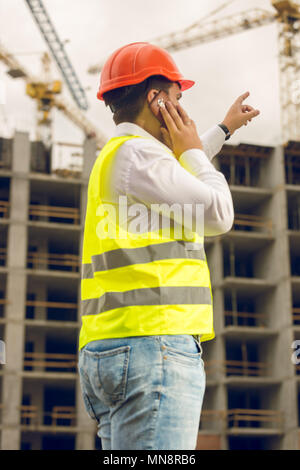 Tonica foto di un uomo in giubbotto di sicurezza e protettivo di compressori hardhat parla al telefono e che puntano a edificio in costruzione. Concetto di investire nel settore immobiliare Foto Stock