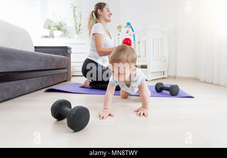 Adorable baby boy strisciando sul pavimento mentre sua madre la pratica fitness in casa Foto Stock