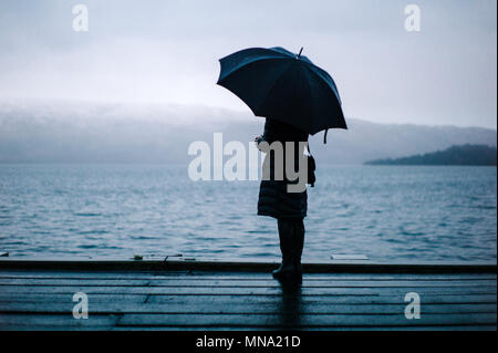 Donna che guarda fuori sopra Loch Lomond con un ombrello in una nebbiosa inverni freddi giorno. Foto Stock