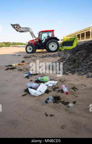 Spazzatura in primo piano su una tranquilla spiaggia di sabbia che si trova al centro turistico presto su un luminoso mattinata estiva essendo pulito dal trattore in background con surf rastrello. Foto Stock