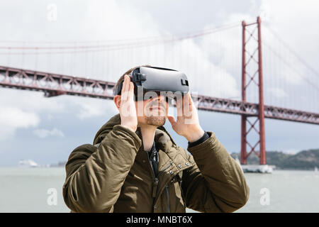 Un uomo utilizza occhiali per realtà virtuale. Xxv Aprile ponte a Lisbona in background. Il concetto di viaggio virtuale. Il concetto di moderne tecnologie e il loro uso nella vita di tutti i giorni Foto Stock