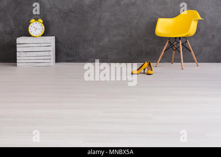 Allarme giallo orologio, sedia, tacchi alti e una scatola di legno in piedi in interni con luce pavimento e muro di cemento Foto Stock
