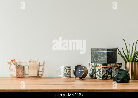 Organizzato un tavolo con scatole floreali, mug, orologio e scatola di metallo su una parete vuota Foto Stock