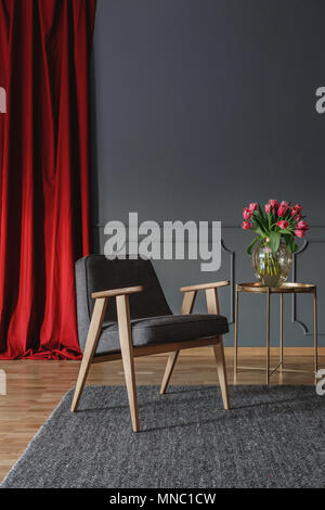 Grigio poltroncina in legno su un tappeto accanto alla tabella di oro in salotto interno con drappi rossi Foto Stock