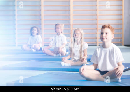 Curioso e stupito i bambini a scuola sportswear seduto in una palestra interno durante attività extra-scolastiche yoga classe e guardando in direzione di un flar Foto Stock