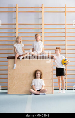 La scuola primaria ragazze seduti sulla parte superiore della scatola di vaulting e un ragazzo tenendo un pallavolo durante una pausa di gioco presso la palestra di classe Foto Stock