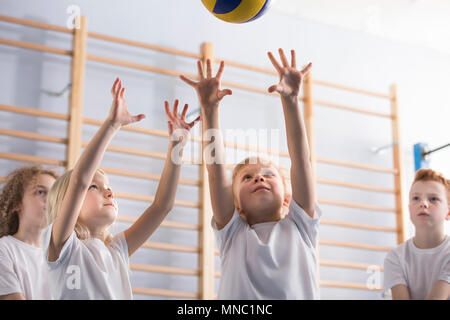 Ragazza giovane con le braccia in alto saltando a colpire un pallavolo durante una partita con la sua scuola compagni di squadra in attività extra-scolastiche educazione fisica classe in Gy Foto Stock