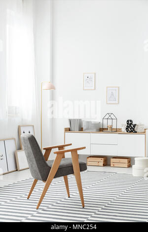 Casse di legno sotto un bianco credenza e una moderna e confortevole poltrona di colore grigio in un elegante salotto interno con un tappeto a strisce e muro bianco Foto Stock