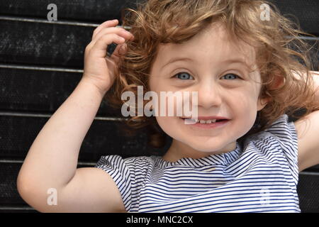 Angelic giovani dai capelli ricci ragazza punti all'interno di lei a bocca aperta con il suo dito seduto contro un nero parete di mattoni nella città di new york Foto Stock