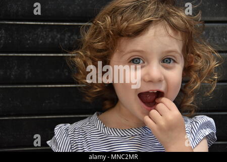 Angelic giovani dai capelli ricci ragazza punti all'interno di lei a bocca aperta con il suo dito seduto contro un nero parete di mattoni nella città di new york Foto Stock