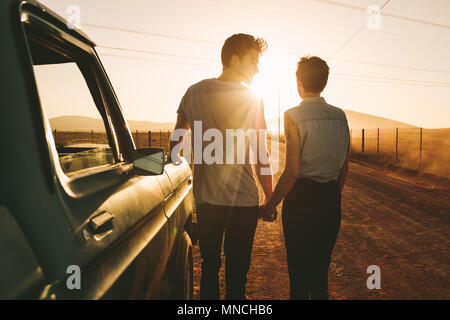 Coppia romantica in piedi tenendo le mani accanto a loro pickup truck durante un viaggio nel paese di lato. Vista posteriore del giovane tenendo le mani e piedi su Foto Stock