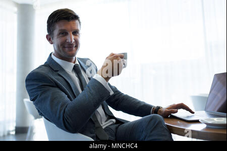 Imprenditore bello sedersi a un tavolo con computer portatile e un caffè. Executive maschio avente una pausa caffè presso l'ufficio. Foto Stock