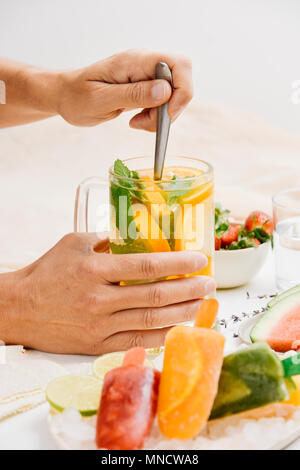 Giovane uomo agitazione un vaso con un drink rinfrescante, con fettine di arancia e menta, collocato su un tavolo accanto a una ciotola con fragole, una piastra con waterm Foto Stock