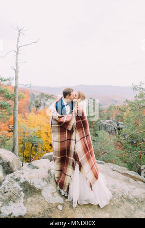 A tutta lunghezza foto del kissing sposa giovane avvolto in maglia plaid. Posizione delle montagne. Foto Stock