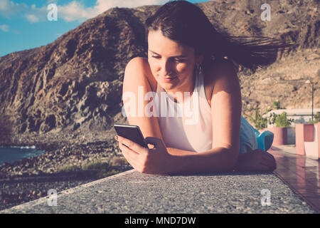 Bellissimi capelli lunghi donna spagnola utilizzare il cellulare per inviare messaggio durante una vacanza a Tenerife. Fissare su un muro vicino alla spiaggia sotto un cielo blu w