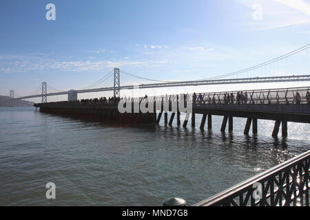 La gente sul molo con Oakland Bay Bridge di tutto il golfo di background, San Francisco, Stati Uniti d'America Foto Stock