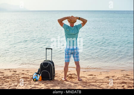 L'uomo turistico in abiti estivi con una valigia in mano, guardando il mare sulla spiaggia, il concetto di tempo per viaggiare Foto Stock