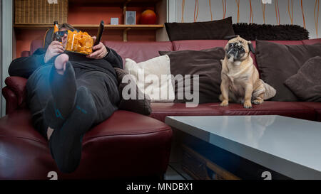 Un uomo mangia chips davanti alla TV. Il suo cane è seduta accanto a lui sul lettino. L'uomo ha un foro in calze e un 3 D bicchieri messo su. Frog p Foto Stock