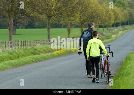 Stanchi dopo il lungo ciclo di corsa, padre e figlio sono a piedi & spingendo le loro biciclette lungo la panoramica strada di campagna - vicino a Ilkley, North Yorkshire, Inghilterra, Regno Unito. Foto Stock